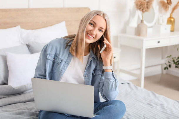 Beautiful young woman with laptop talking by mobile phone at home - Foto, Bild
