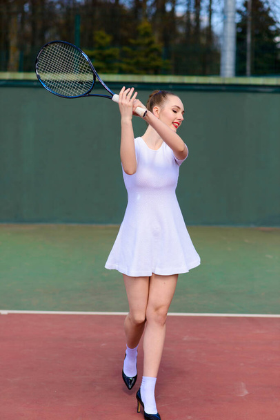 Sahada tenis raketi tutan beyaz elbiseli ve topuklu ayakkabılı seksi kız tenisçi. Genç kadın tenis oynuyor. - Fotoğraf, Görsel