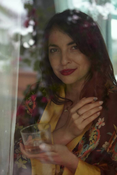 Woman Smoking THC CBD Marijuana Joint in Window in vintage robe with glass of wine. She is separated from the photographer by the window. - Photo, image