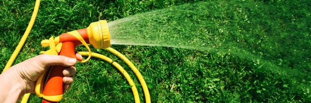 Hand houdt gele slang met mondstuk op de achtergrond van gras tuin met spatten - Foto, afbeelding