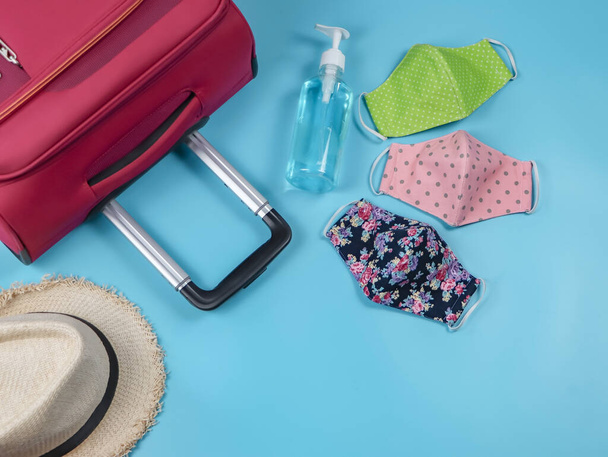 COVID-19 Prävention auf Reisen und neues normales Lebensstilkonzept. Draufsicht auf drei bunte Stoffgesichtsmasken, Alkohol-Händedesinfektionsgel, Hut und Koffer auf blauem Hintergrund. - Foto, Bild