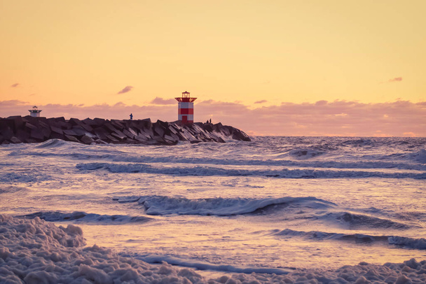 Красно-белый полосатый маяк во время яркого заката. Вода бьет по черным валунам волнами. Драматическое небо в золотом цвете. Шевенхель, Нидерланды
 - Фото, изображение