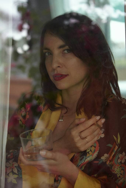 Woman Smoking THC CBD Marijuana Joint in Window in vintage robe with glass of wine. She is separated from the photographer by the window. - Fotó, kép