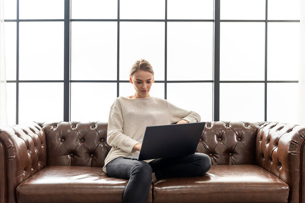 Krásná žena pracuje doma se svým laptopem. Ta žena sedí na hnědé kožené pohovce. - Fotografie, Obrázek