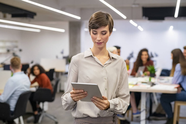 Happy young businesswoman using digital tablet in modern open office. Business woman typing on digital tablet with colleague working in background. Portrait of latin woman standing in a coworking space while working on tablet. - Fotó, kép