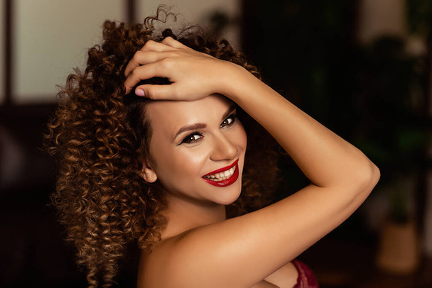 Portrait of curly and sunburnt european woman in a dark flat, happy stylish smiling caucasian model, woman laughs and looks at you, pretty young woman with a red lips, girl straightens her hair - Foto, imagen