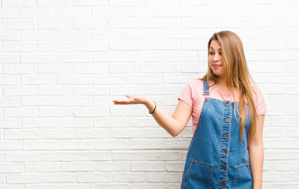 young blonde woman feeling happy and smiling casually, looking to an object or concept held on the hand on the side against brick wall - Foto, immagini