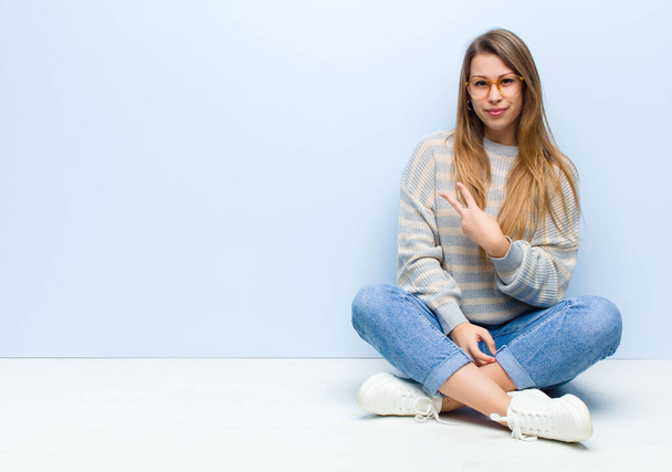 young blonde woman feeling happy, positive and successful, with hand making v shape over chest, showing victory or peace sitting on the floor - 写真・画像