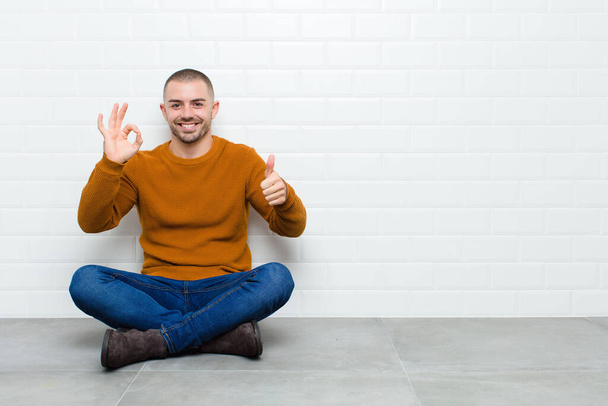 young handsome man feeling happy, amazed, satisfied and surprised, showing okay and thumbs up gestures, smiling sitting on the floor - Фото, зображення