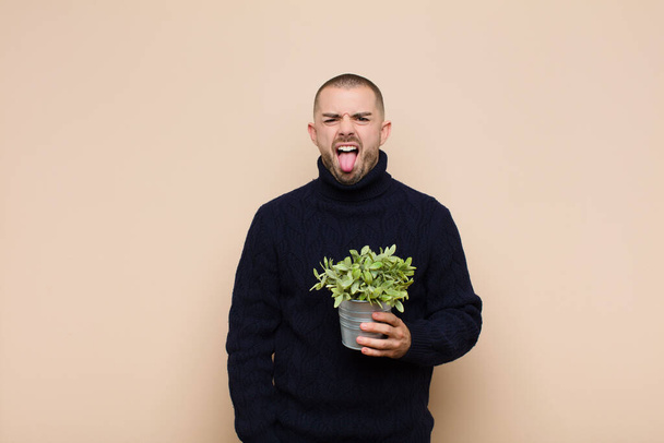 young handsome man feeling disgusted and irritated, sticking tongue out, disliking something nasty and yucky holding a plant - Fotoğraf, Görsel