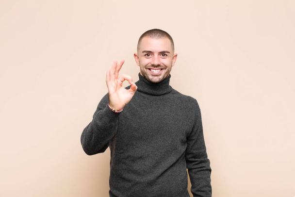 young handsome man feeling happy, relaxed and satisfied, showing approval with okay gesture, smiling against flat wall - Zdjęcie, obraz