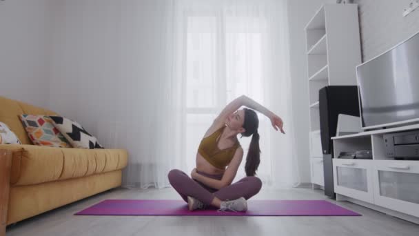 Joven hermosa mujer está practicando estiramiento en estera violeta en casa
 - Metraje, vídeo