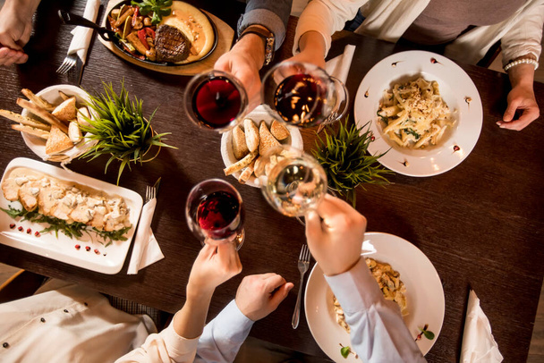 Τέσσερα χέρια με κόκκινο κρασί που ψήνεται πάνω από σερβίρεται τραπέζι με φαγητό στο εστιατόριο - Φωτογραφία, εικόνα