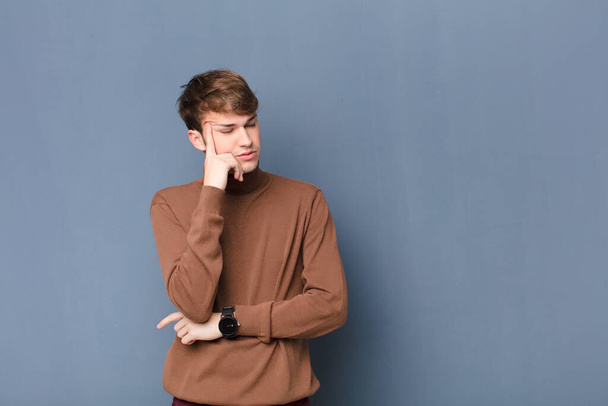 young blonde man with a concentrated look, wondering with a doubtful expression, looking up and to the side isolated against flat wall - Foto, Bild