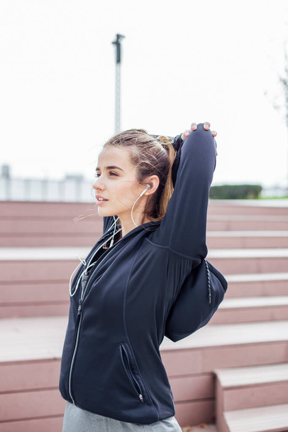 Fit young woman with earphones doing stretching exercises on urban stairs outdoors - Фото, зображення