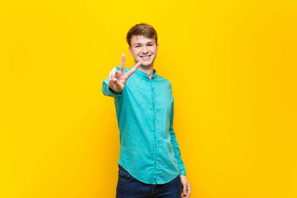 young blonde man smiling and looking happy, carefree and positive, gesturing victory or peace with one hand isolated against flat wall - Photo, image