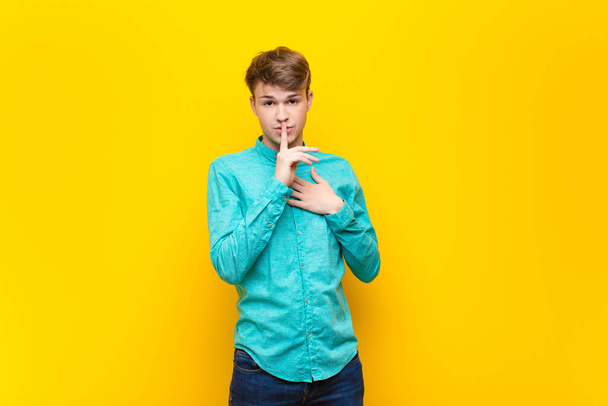 young blonde man looking serious and cross with finger pressed to lips demanding silence or quiet, keeping a secret isolated against flat wall - Foto, afbeelding