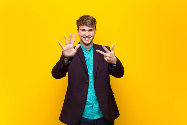 young blonde man smiling and looking friendly, showing number eight or eighth with hand forward, counting down isolated against flat wall - Fotoğraf, Görsel