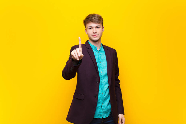 young blonde man smiling and looking friendly, showing number one or first with hand forward, counting down isolated against flat wall - Fotoğraf, Görsel