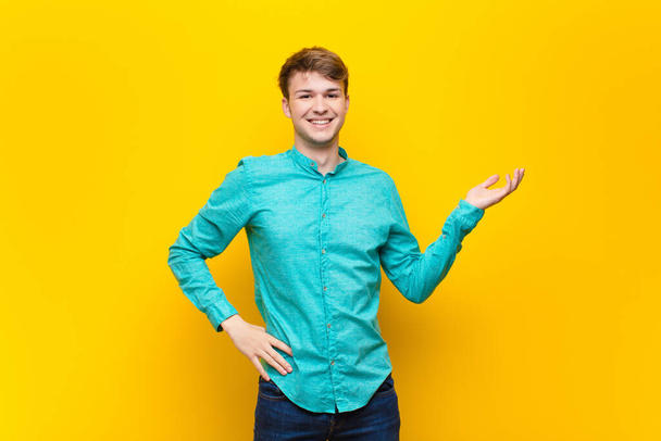 young blonde man smiling, feeling confident, successful and happy, showing concept or idea on copy space on the side isolated against flat wall - Photo, image
