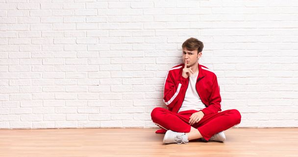young blonde man asking for silence and quiet, gesturing with finger in front of mouth, saying shh or keeping a secret sitting on the floor - Photo, Image