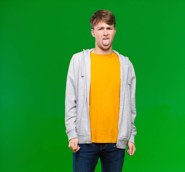 young blonde man feeling disgusted and irritated, sticking tongue out, disliking something nasty and yucky against chroma key wall - Fotoğraf, Görsel