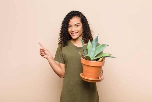 young black woman smiling cheerfully, feeling happy and pointing to the side and upwards, showing object in copy space holding a cactus - Foto, imagen