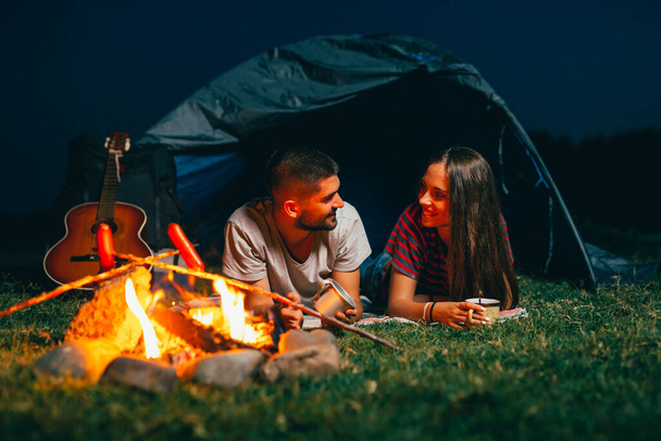 romantic couple on night camping, enjoying company and barbecue sausages on camping fire - Photo, Image