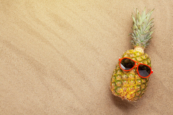 Ripe pineapple with sunglasses over sand background with copy space. Travel and vacation concept. Top view flat lay - Photo, Image