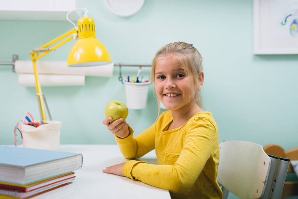 schoolgirl in her room sitting table holding apple - Foto, Imagem