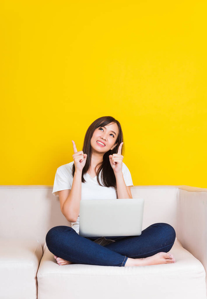 Portrait Asian of happy beautiful young woman work from home she sitting on sofa using laptop computer in house living room pointing finger up away studio shot isolated on yellow background - Photo, Image