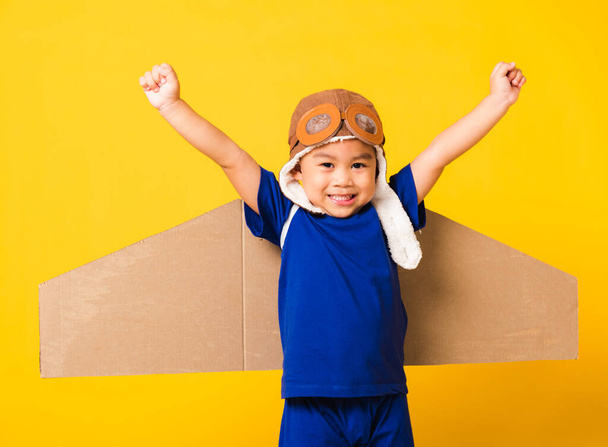 Happy Asian handsome funny child or kid little boy smile wear pilot hat play and goggles raise hand up with toy cardboard airplane wings flying, studio shot isolated yellow background, Startup freedom - Fotó, kép