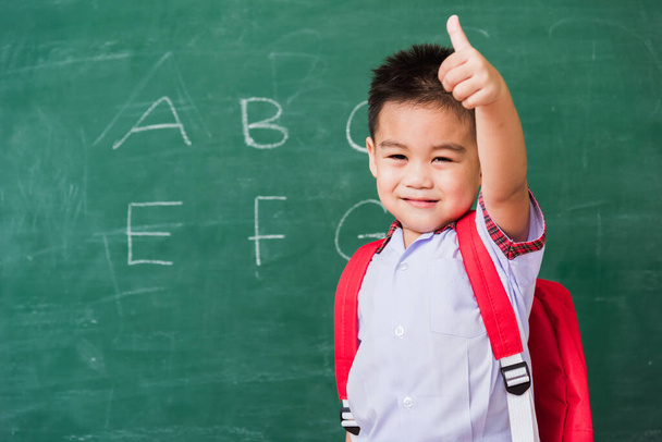 Vissza az iskolába. Boldog ázsiai vicces aranyos kisfiú óvoda diák egyenruha iskolatáska mosolygós show ujj fel a zöld iskola táblán, Az első alkalom, hogy az iskolai oktatási koncepció - Fotó, kép