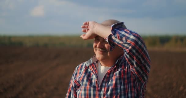 Un granjero mayor en un campo mirando a la distancia. Agricultor senior de pie en el campo de soja examinando cultivos al atardecer
 - Imágenes, Vídeo