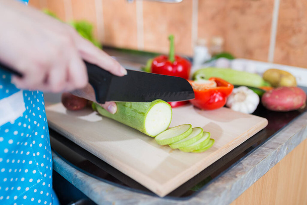 Close up of woman in apron slicing zucchini on cutting board in kitchen. Healthy meal preparation concept. - Foto, imagen