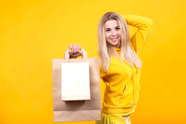 Portret młodej pięknej białej blondynki z papierowymi eko-torbami w żółtym sportowym garniturze, odizolowany na żółtym tle, uśmiechnięty, patrzący w kamerę. - Zdjęcie, obraz