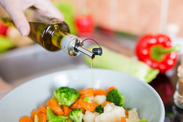 Close up of housewife holding bottle of olive oil and cooking pan with various vegetables in kitchen. - Foto, Bild