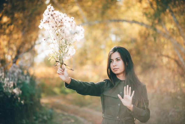 Dreamy beautiful girl with thistle flowers bouquet in golden sunlight on bokeh background with yellow leaves. Inspired girl enjoys sunset in autumn forest. Female beauty portrait among autumn foliage. - Photo, Image