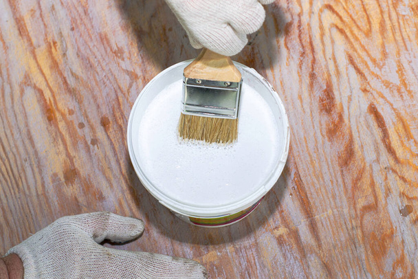 İş eldivenli bir adam beyaz boyayla ahşap bir yüzey boyuyor, boya fırçasını boya kavanozuna koyuyor. - Fotoğraf, Görsel
