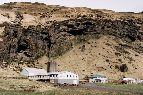 Mezőgazdasági vállalkozások lakóházai és termelőlétesítményei, az izlandi hegy lábánál. Legeltetésre szánt zöld rét. - Fotó, kép