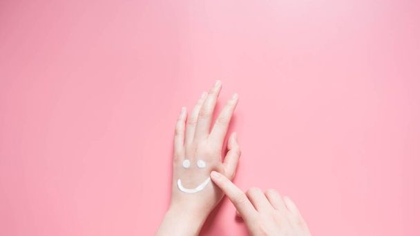 Hermosas manos de mujer aplicando crema hidratante sobre fondo rosa. Sonriente forma emoji creado a partir de la loción. Cuida tu piel corporal. Concepto de salud y belleza. Vista superior. De cerca.
. - Foto, Imagen