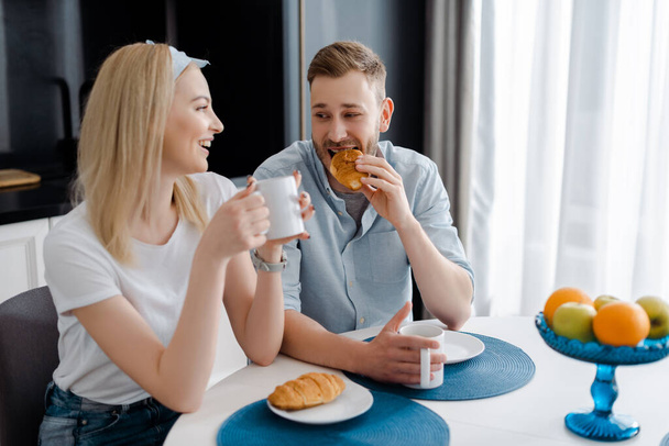 happy girl holding cup and looking at boyfriend eating croissant  - Foto, Bild