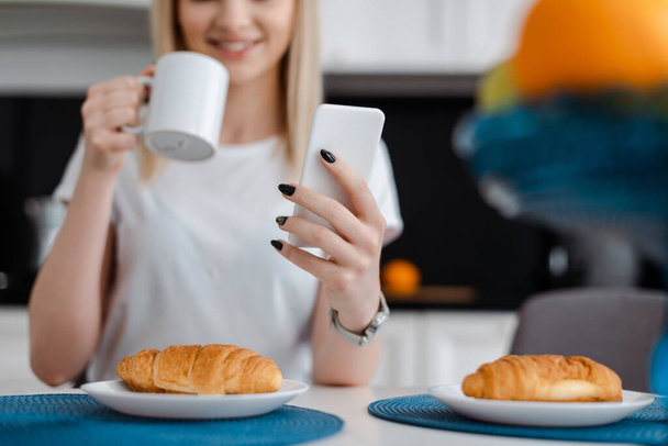 cropped view of girl holding smartphone and cup near croissants  - Фото, зображення