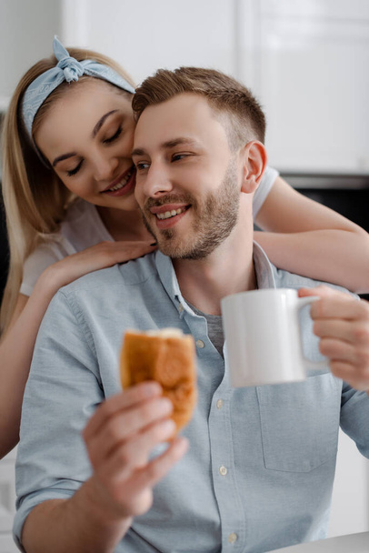 selective focus of girl touching happy boyfriend holding cup of coffee and croissant in kitchen  - Photo, Image