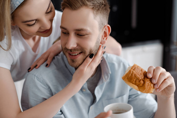 selective focus of beautiful girl touching handsome boyfriend holding cup of coffee and croissant in kitchen  - Photo, Image