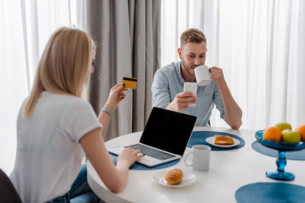 selektiver Fokus der Frau mit Kreditkarte in der Nähe von Laptop mit leerem Bildschirm und hübschem Freund, der Kaffee trinkt, während er Smartphone benutzt  - Foto, Bild