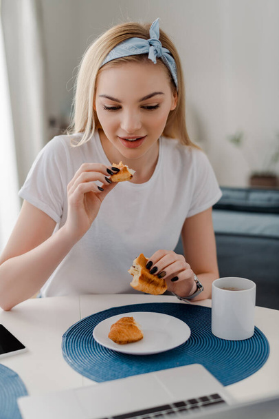 selective focus of attractive freelancer holding croissant near laptop  - Foto, afbeelding