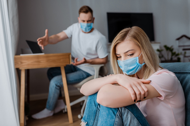 selective focus of girl in medical mask sitting near angry boyfriend gesturing while quarreling at home  - Фото, изображение