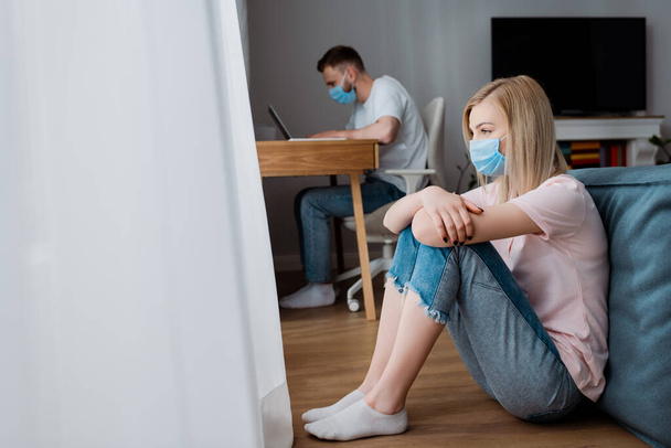 foco seletivo da menina em máscara médica sentado no chão perto do namorado freelancer trabalhando em casa
  - Foto, Imagem