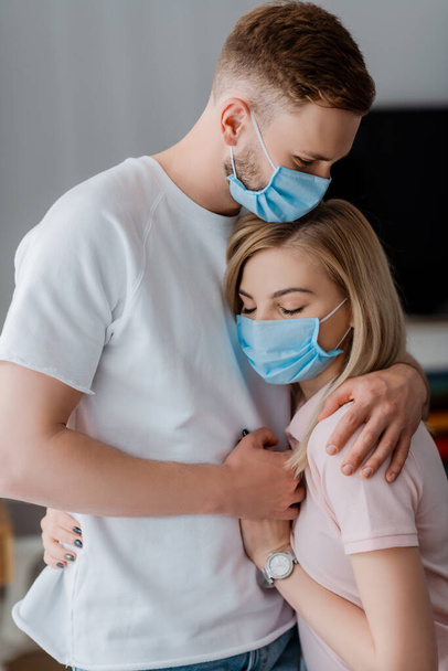 pareja en máscaras médicas cogidas de la mano y abrazándose en casa
  - Foto, Imagen